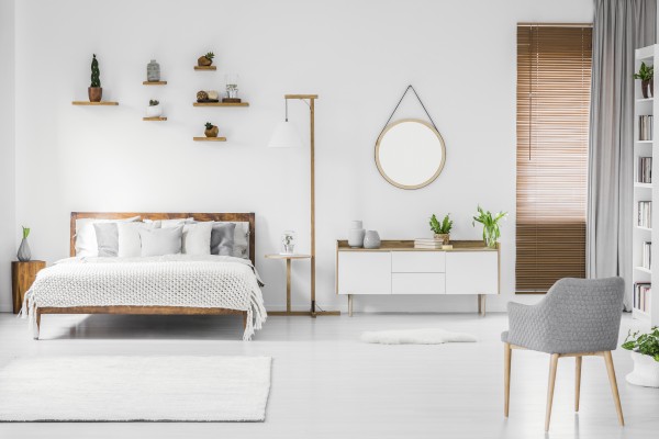Spacious designer white bedroom interior with wooden bed with bedding and pillows, night-table, small shelves above and modern armchair on the right. Real photo.